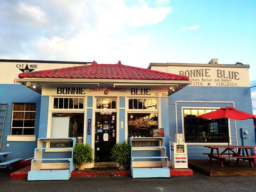 The charming exterior of Bonnie Blue Southern Market and Bakery in Winchester, Virginia, featuring a light blue building with a distinctive red tiled roof, large windows displaying menu items, and outdoor seating with a red umbrella, presenting a local eatery as one of the appealing things to do in Winchester VA.