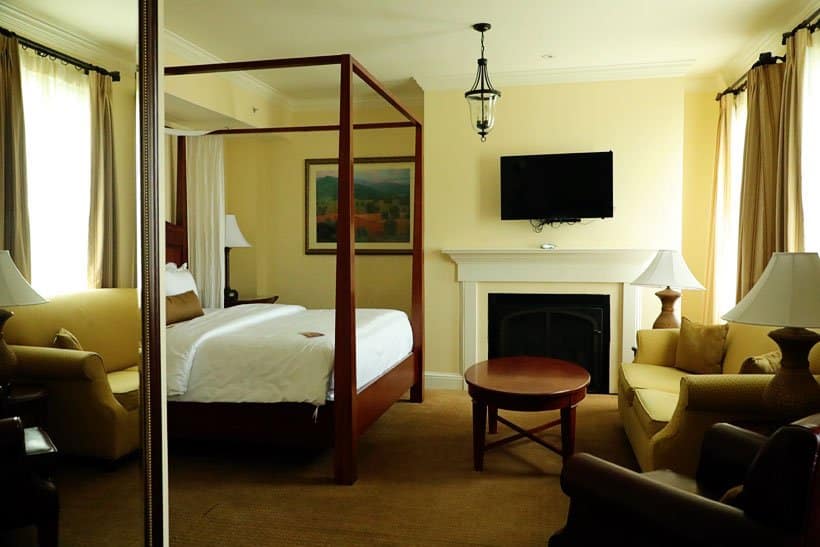 A well-appointed hotel room at The George Washington A Wyndham Grand Hotel in Winchester, VA, featuring a four-poster bed with white linens, a fireplace with a mounted television above it, comfortable seating including a sofa and armchairs, and elegant lamps, showcasing a luxurious accommodation option for things to do in Winchester VA.