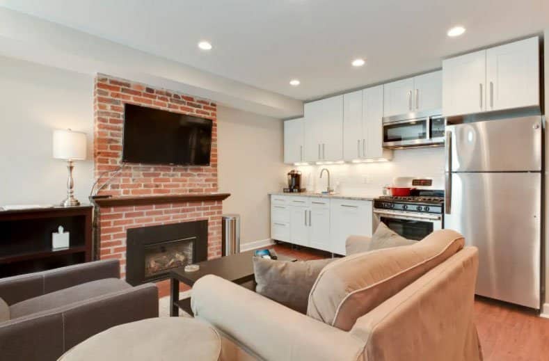 21 Extraordinary Airbnb Washington DC Rentals to Book Now - A small, open-plan living space featuring a gray sofa, a tan armchair, a coffee table, and a brick fireplace with a mounted television. Adjacent to the living area is a compact kitchen with white cabinets and stainless steel appliances.