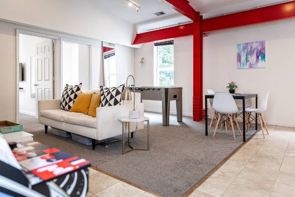 21 Extraordinary Airbnb Washington DC Rentals to Book Now - A modern living space with a white sofa accented by patterned and mustard-colored pillows, a small round side table with a lamp, a foosball table, and a small black dining table with white chairs. Red structural beams are visible on the ceiling and wall.
