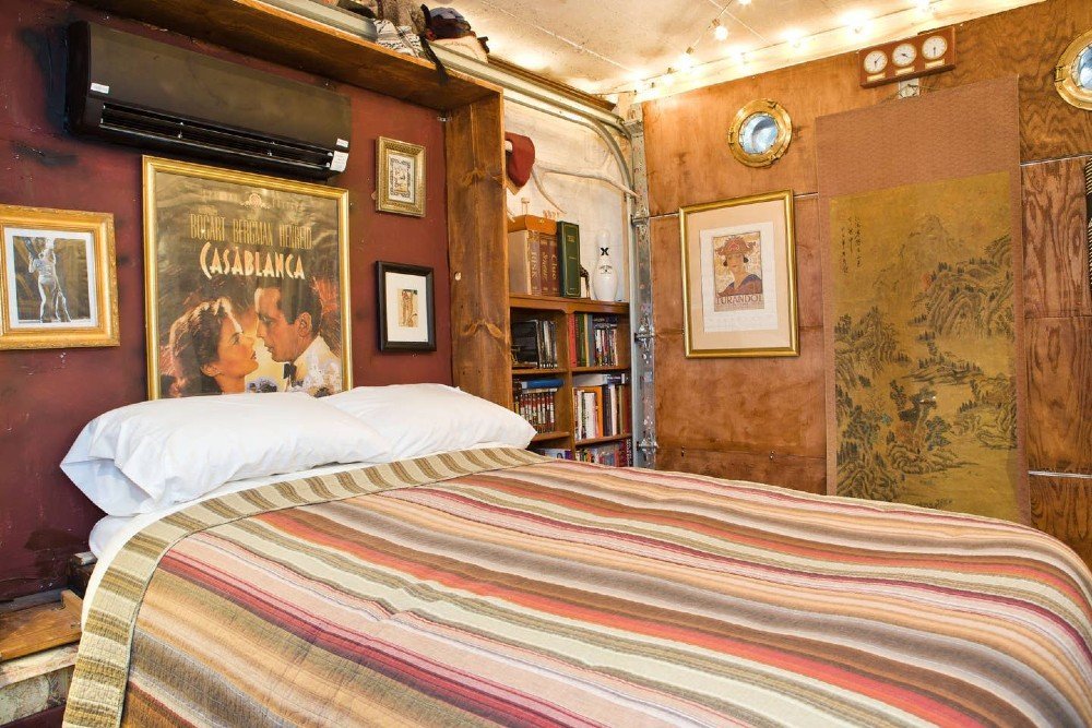 21 Extraordinary Airbnb Washington DC Rentals to Book Now - A bedroom space featuring a bed with a multi-colored striped blanket, a "Casablanca" movie poster in a gold frame, a small air conditioning unit, and shelves filled with books.