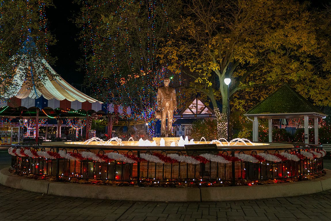 A festive night view of Hersheypark at Christmas, with a statue in the center of a fountain surrounded by colorful lights.