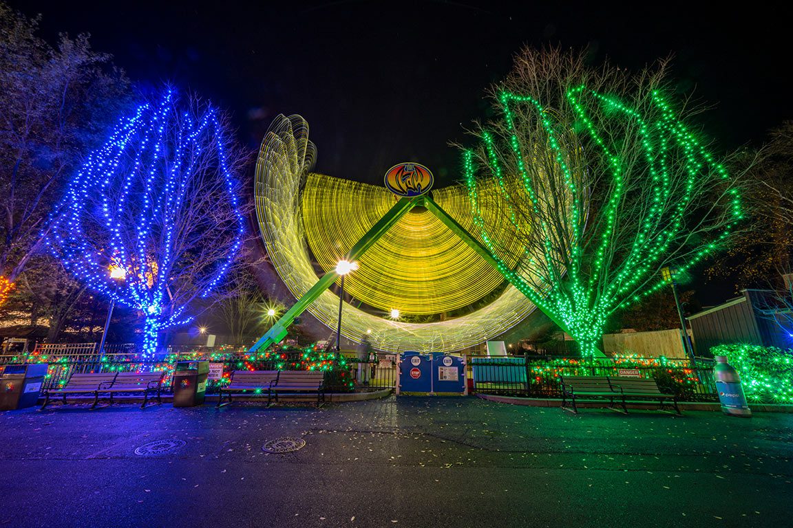 Vibrant holiday lights illuminate trees and a spinning ride at Hersheypark at Christmas, creating a festive and colorful night display.