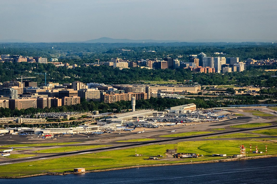 An aerial view shows Ronald Reagan Washington National Airport (DCA) situated along the Potomac River, with the urban landscape of Arlington, VA, visible in the background, featuring various buildings and green spaces extending towards the horizon. While the airport itself is a transportation hub, exploring the nearby attractions is one of the many things to do in Arlington VA after arriving.