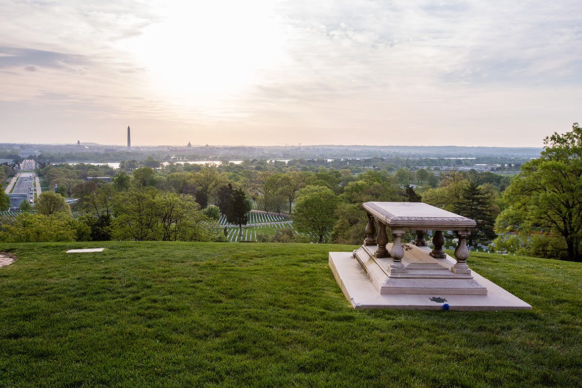 A serene landscape view from Arlington National Cemetery, featuring the tomb of an unknown soldier in the foreground on a grassy hill. Rows of white headstones stretch across the rolling green landscape, leading towards a distant view of Washington D.C. landmarks under a soft, cloudy sky. This poignant scene represents just one of the many historical things to do in Arlington VA.
