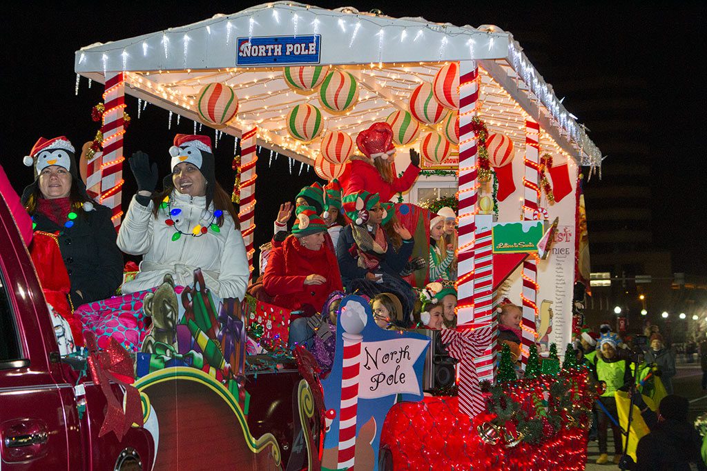 People on a festive North Pole-themed float, adorned with Christmas lights and decorations, as part of a holiday parade in Hampton Roads. The float is surrounded by festive red and green colors, with children and adults dressed in winter attire, enjoying the parade. Christmas Lights in Hampton Roads create a magical atmosphere during the holiday season.