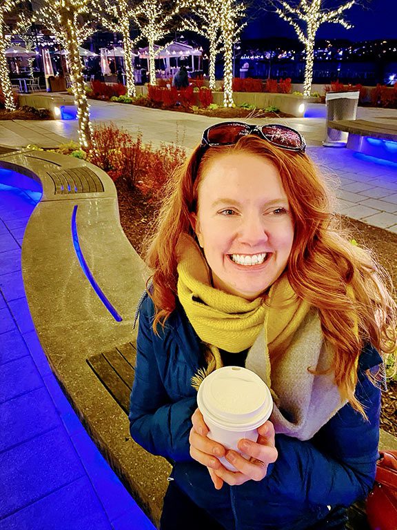 A smiling woman, cozy in the winter season, holds a steaming hot chocolate, its warmth visible in the crisp air. This inviting scene captures the delight of finding the best hot chocolate near me or the best hot chocolate in dc.