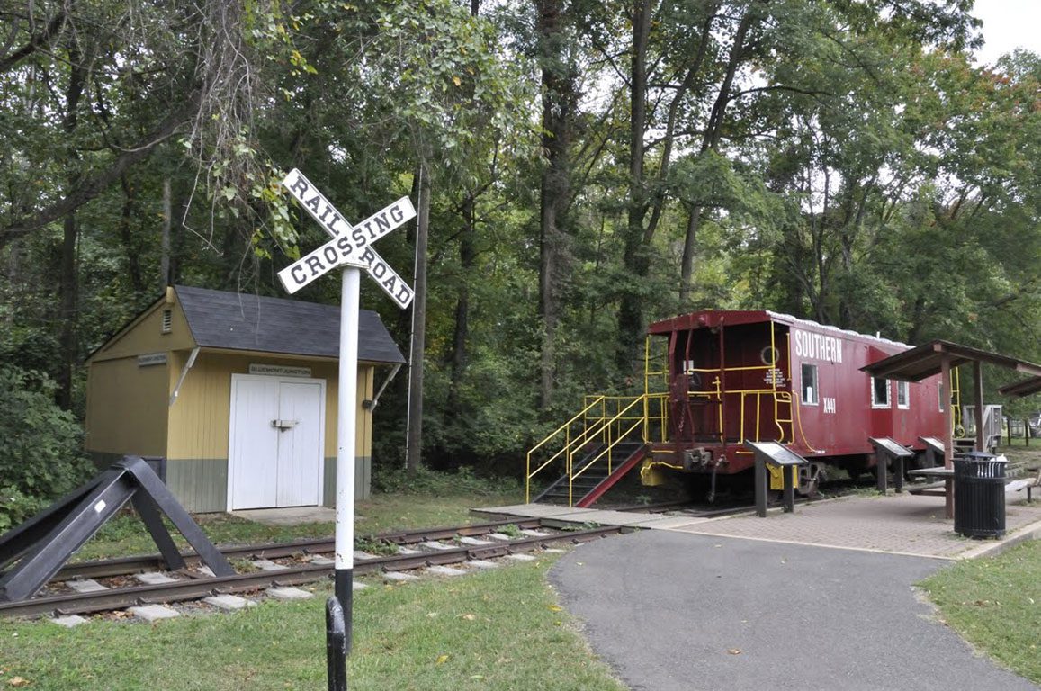 An outdoor exhibit featuring a red Southern Railway caboose with yellow railings, parked on a section of railroad tracks next to a small yellow and white building and a "RAILROAD CROSSING" sign. This historical display, set amongst trees, offers a glimpse into the area's past and is one of the unique things to do in Arlington VA.