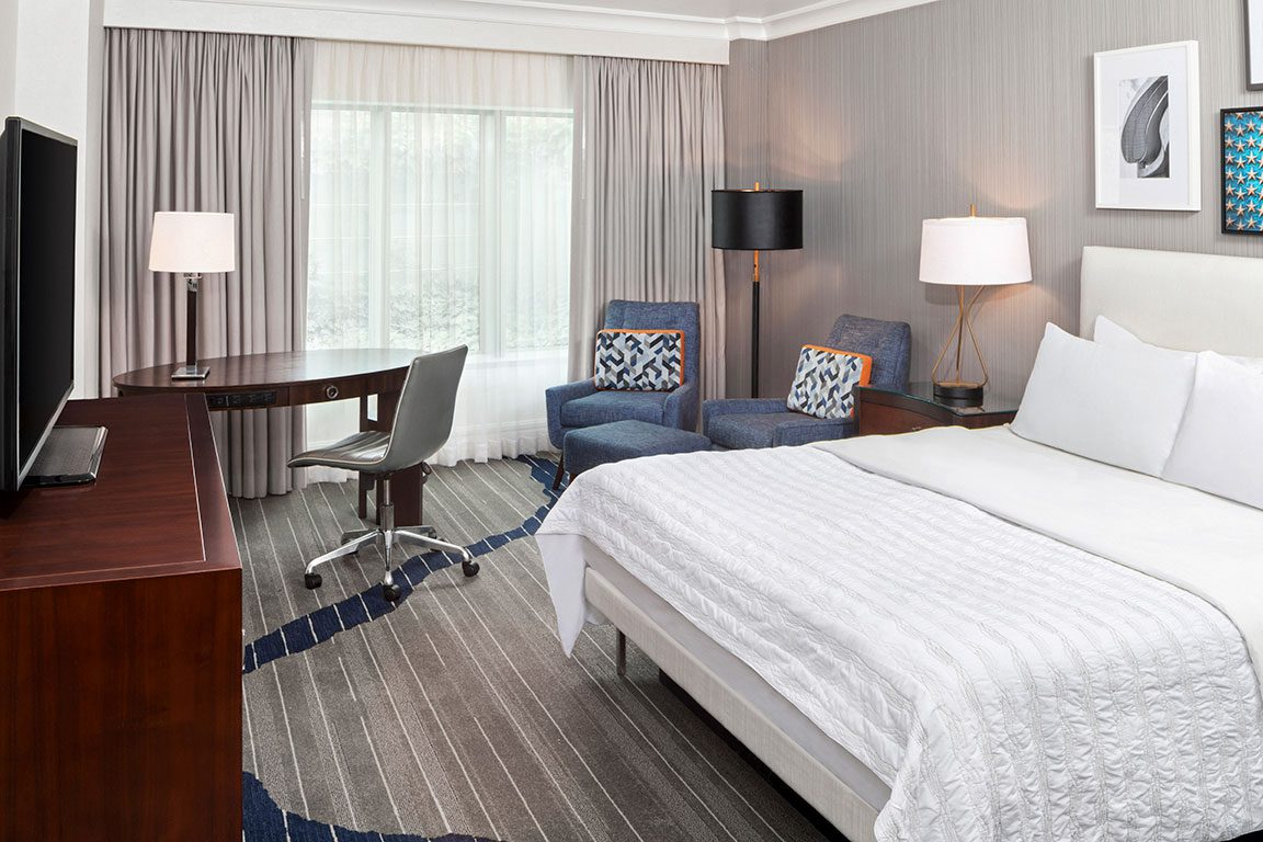 A well-appointed hotel room at what might be Le Méridien Arlington features a comfortable bed with white linens, two blue armchairs with patterned pillows, a dark wood desk and chair, and a flat-screen TV. The room is decorated in neutral tones with pops of blue and orange. Staying in a hotel like this provides a convenient base for exploring the many things to do in Arlington VA.