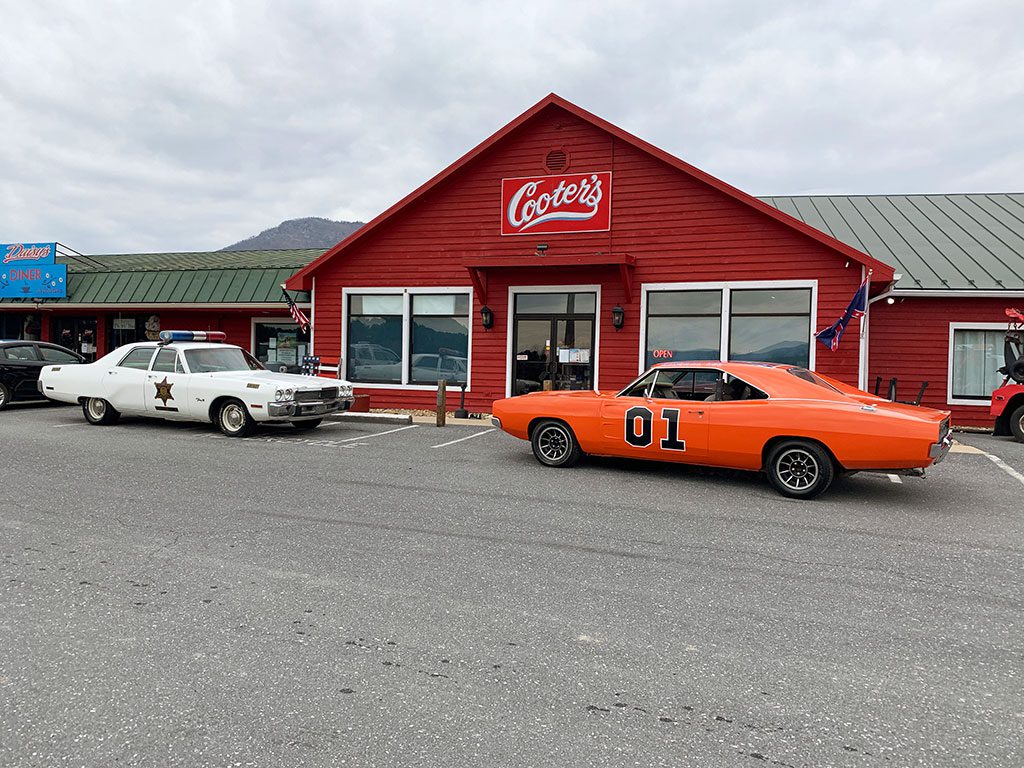 A daytime view of "Cooter's" in Luray, VA, a red building with the business's name in white script above the entrance. Parked in the asphalt parking lot in front are two iconic vehicles: a white police car with a star emblem on the door and the orange "General Lee" car with the number "01" prominently displayed on its side. This suggests a "Dukes of Hazzard" themed attraction. Visiting Cooter's is one of the nostalgic things to do in Luray VA.