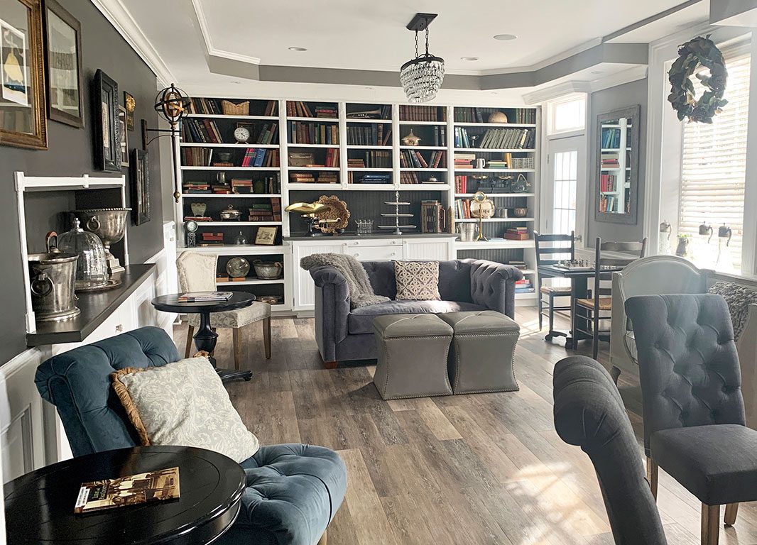 An interior view of a cozy and stylishly decorated room, possibly a common area or seating lounge at Hotel Laurance in Luray, VA. The room features a large built-in bookcase filled with books and decorative items, comfortable seating including a tufted gray loveseat with ottomans and a blue velvet armchair, and small round tables. The decor includes a chandelier, artwork on the walls, and a wreath on the window. Relaxing in a charming space like this is one of the pleasant things to do in Luray VA while visiting or staying at local establishments.