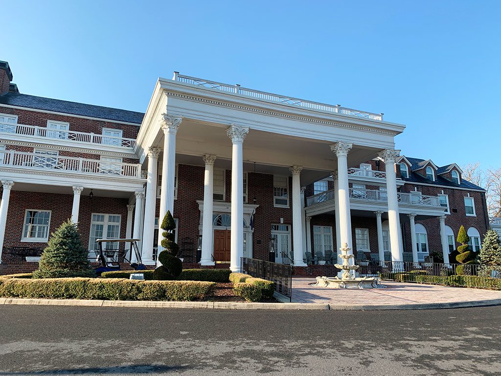 A daytime, eye-level view of the Mimslyn Inn, a grand, historic brick building with a prominent white portico supported by large classical columns. The inn features multiple floors with balconies and numerous windows. Manicured hedges, trees, and a stone fountain adorn the front grounds, and an asphalt driveway leads up to the entrance. Staying at the Mimslyn Inn, a notable landmark, is one of the elegant things to do in Luray VA.