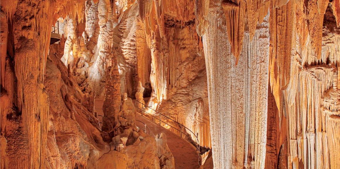 An interior view of the Luray Caverns, featuring impressive geological formations such as stalactites hanging from the ceiling and stalagmites rising from the ground, illuminated with warm lighting. A winding walkway with railings allows visitors to explore the underground landscape. Visiting stunning caverns like this is one of the memorable things to do in Luray VA.