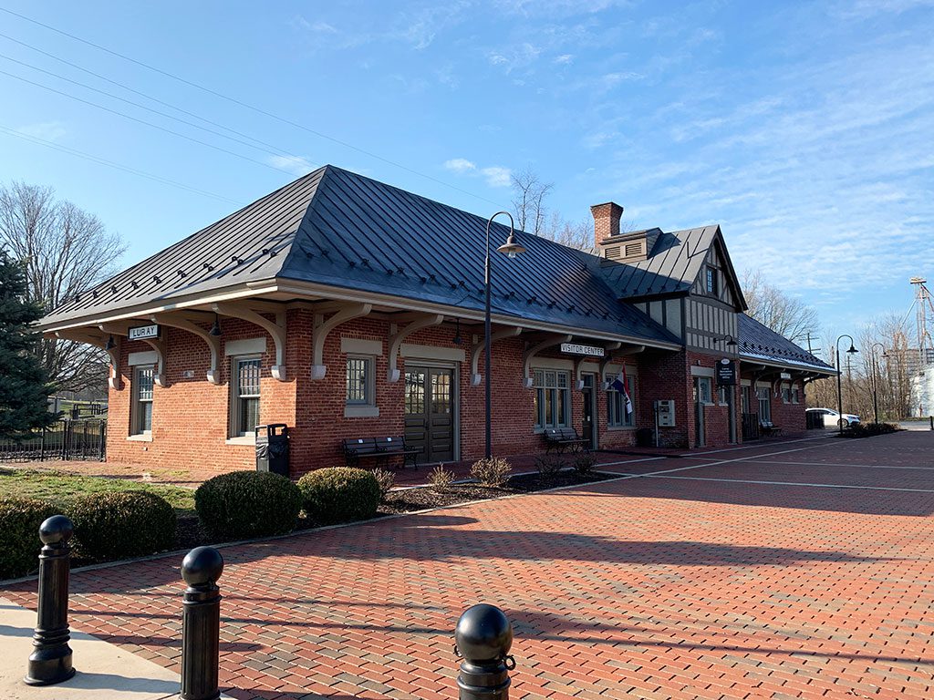 A daytime, eye-level view of the historic Luray Norfolk & Western Passenger Station, a red brick building with a distinctive dark metal roof and decorative brickwork. Signage indicates "LURAY" on one end and "FREIGHT STATION" along the side. The building is situated on a brick-paved area with manicured greenery and black bollards in the foreground. Visiting historical landmarks like the Luray station is one of the interesting things to do in Luray VA.