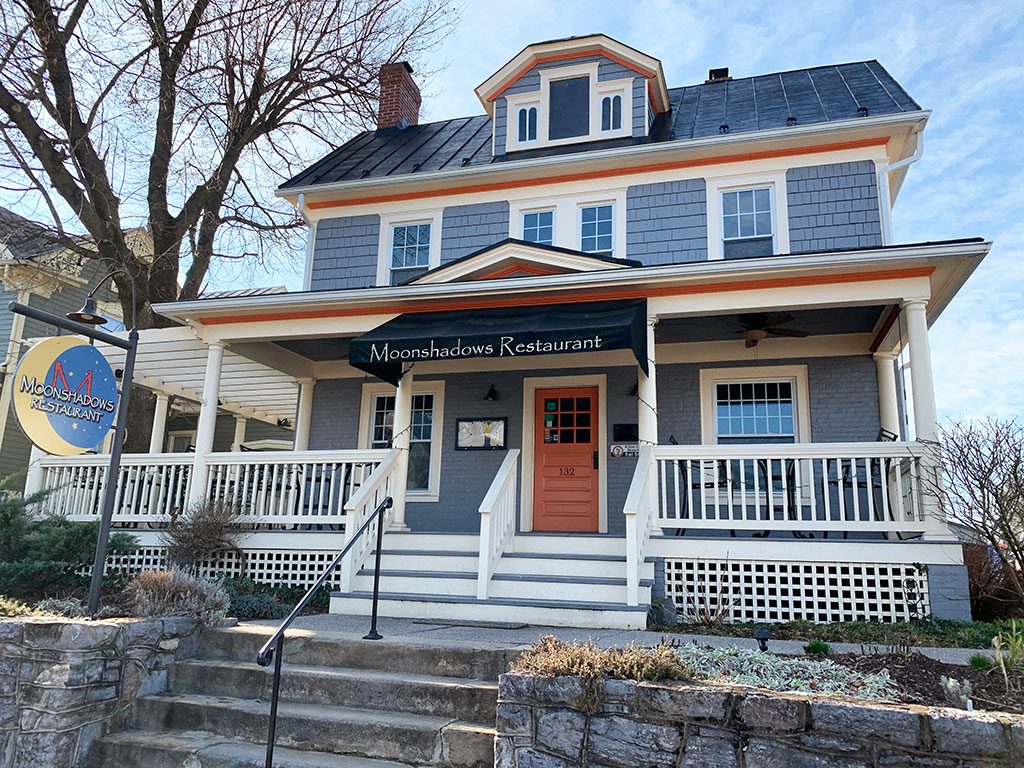 A daytime view of Moonshadows Restaurant, a charming two-story building with gray siding, white trim, and an orange front door. A covered porch with white railings extends across the front, featuring a black awning with "Moonshadows Restaurant" written in white. A round, blue and yellow sign with the restaurant's name hangs near the entrance. Stone steps with a black railing lead up to the porch. Dining at local restaurants like Moonshadows is one of the enjoyable things to do in Luray VA.