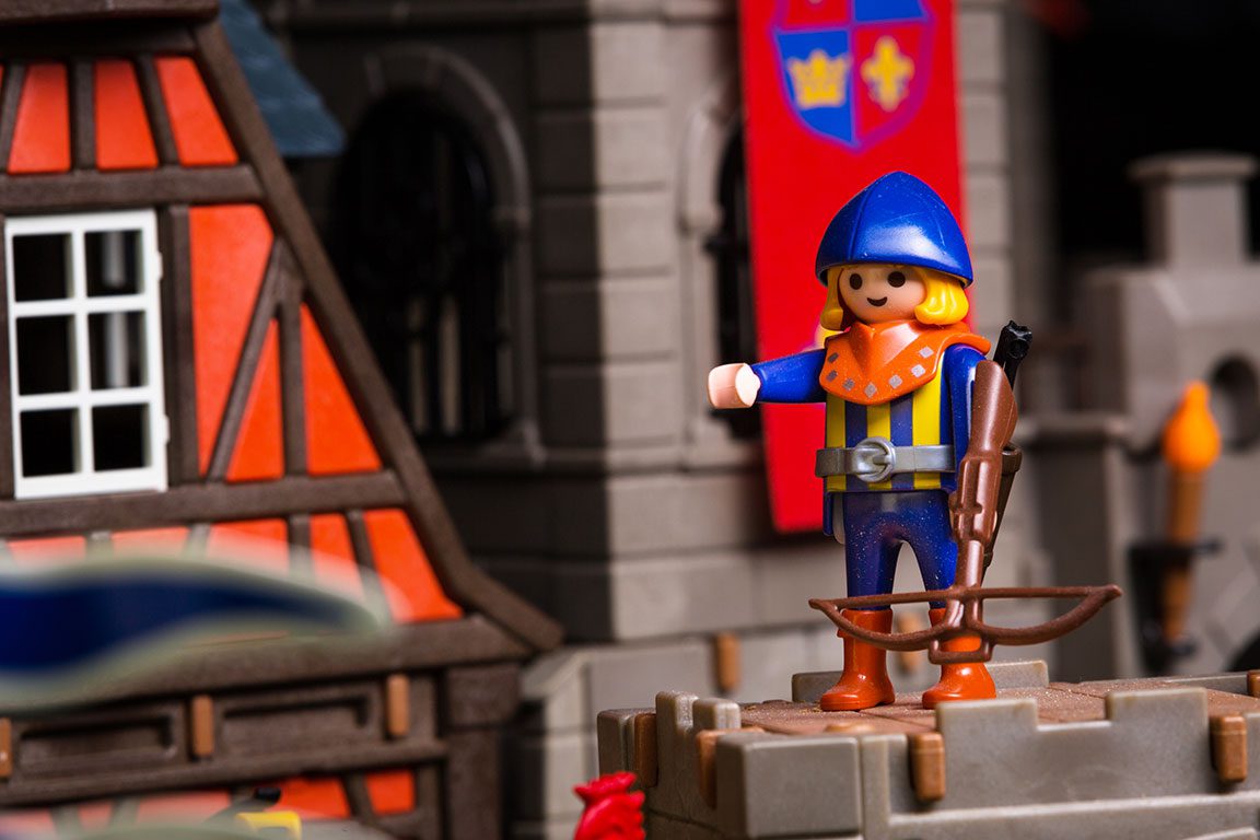 A close-up shot of a Playmobil figure dressed as a medieval archer, likely part of a display at Toy Town Junction. The figure wears a bright blue helmet, an orange and yellow tunic, blue pants, and brown boots. A crossbow is positioned in front of the figure, resting on a gray, crenelated castle wall. In the blurred background, a portion of a brown and orange timber-framed building with white windows is visible on the left, and a gray castle structure with a red banner displaying yellow symbols is on the right. Visiting Toy Town Junction could be one of the playful things to do in Luray VA.