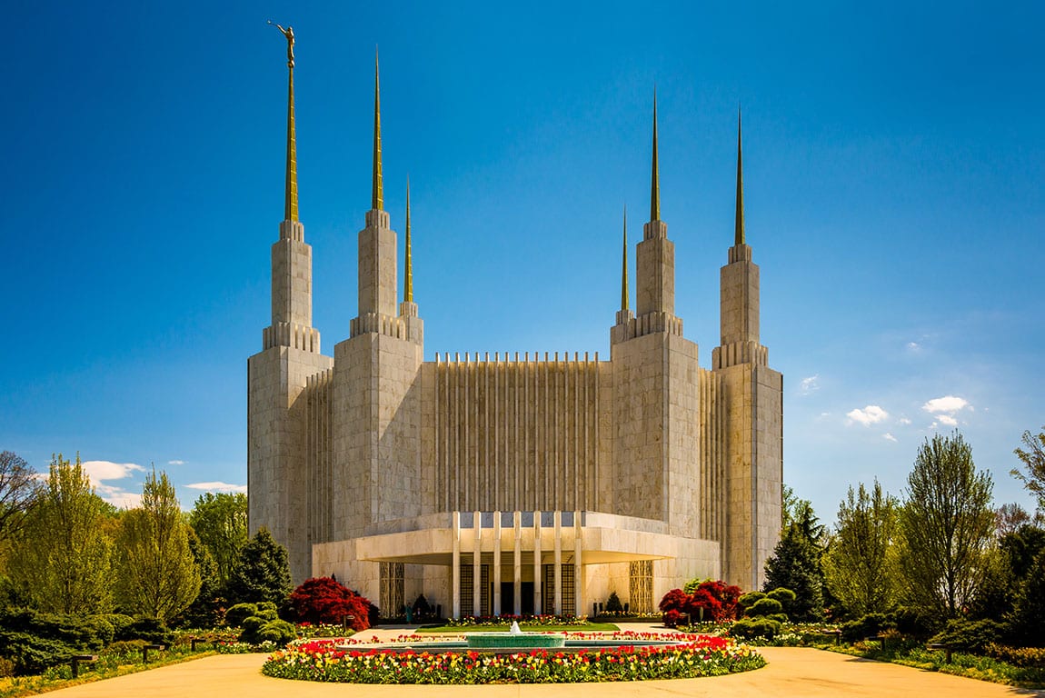 A daytime view of the Washington D.C. Temple of The Church of Jesus Christ of Latter-day Saints, located in Kensington, Maryland, near Silver Spring. The large, white marble temple features six spires, with the tallest one adorned with a golden Angel Moroni statue. A reflecting pool and colorful flowerbeds are in the foreground, along with a paved walkway and surrounding greenery. While visiting the temple grounds is typically for religious purposes, admiring its architecture and serene setting is one of the points of interest near Silver Spring, MD.