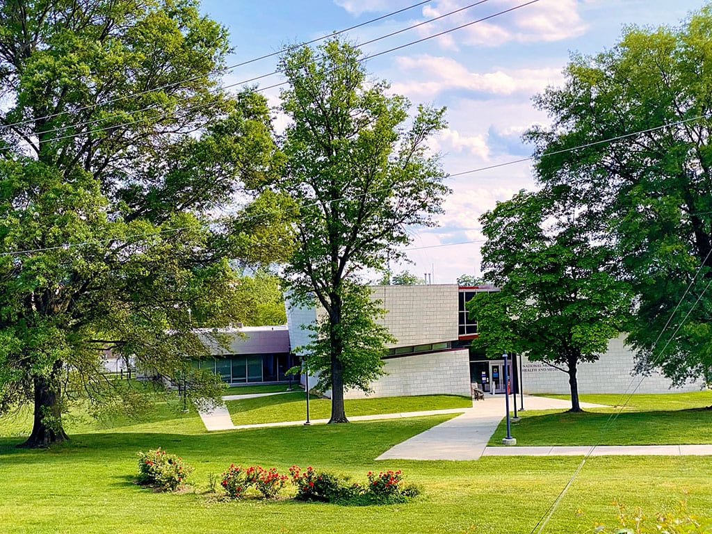 A daytime view of the National Museum Of Health And Medicine. Large trees with green foliage partially obscure the view of the building. A well-maintained green lawn extends in front of the museum. Visiting the National Museum Of Health And Medicine could be one of the educational things to do in Silver Spring MD, although the exact location needs to be verified.