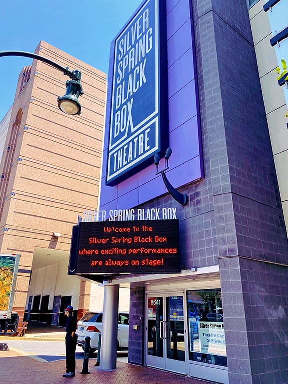 A daytime view of the Silver Spring Black Box Theatre. The building has a modern facade with a large vertical sign displaying the theater's name in white letters on a blue and purple background. An electronic marquee below welcomes visitors and announces "exciting performances are always on stage!" A person stands on the sidewalk near the entrance. Attending a performance at the Silver Spring Black Box Theatre is one of the cultural things to do in Silver Spring MD.