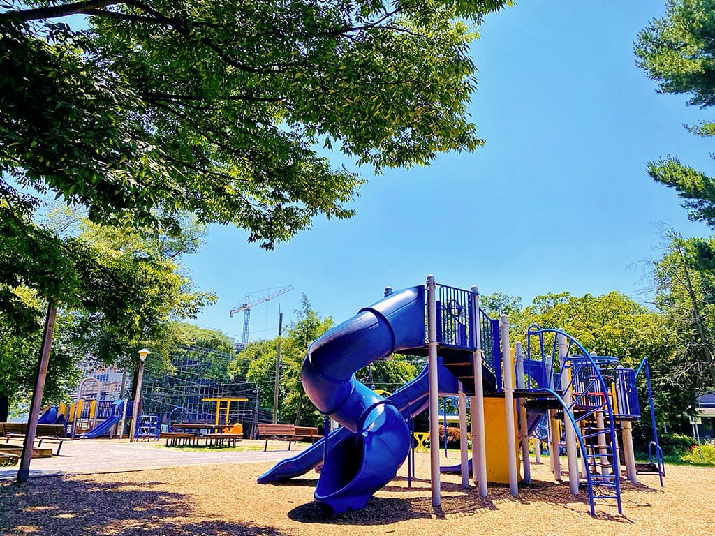A daytime view of a colorful playground at what might be Woodside Urban Park in Silver Spring, MD. The playground features a large blue spiral slide, other slides, climbing structures, and swings, all set on a mulch ground covering. Mature green trees surround the play area, casting dappled shade. Benches and picnic tables are visible in the background. Spending time at a local park like this is one of the recreational things to do in Silver Spring MD.