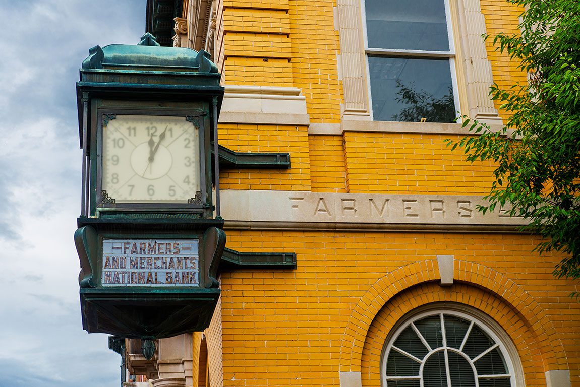 A close-up of the architectural details of the Farmers and Merchants National Bank building in Winchester, Virginia, featuring a vintage street clock displaying the time, the bank's name etched in stone, and the building's yellow brick facade, highlighting a historical landmark and a potential point of interest for things to do in Winchester VA.
