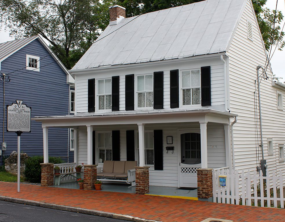 A charming two-story white clapboard house with black shutters and a gray metal roof, featuring a welcoming front porch with brick supports and a swing, identified by a historical marker as the Patsy Cline Historic House in Winchester, Virginia, a notable landmark for things to do in Winchester VA.