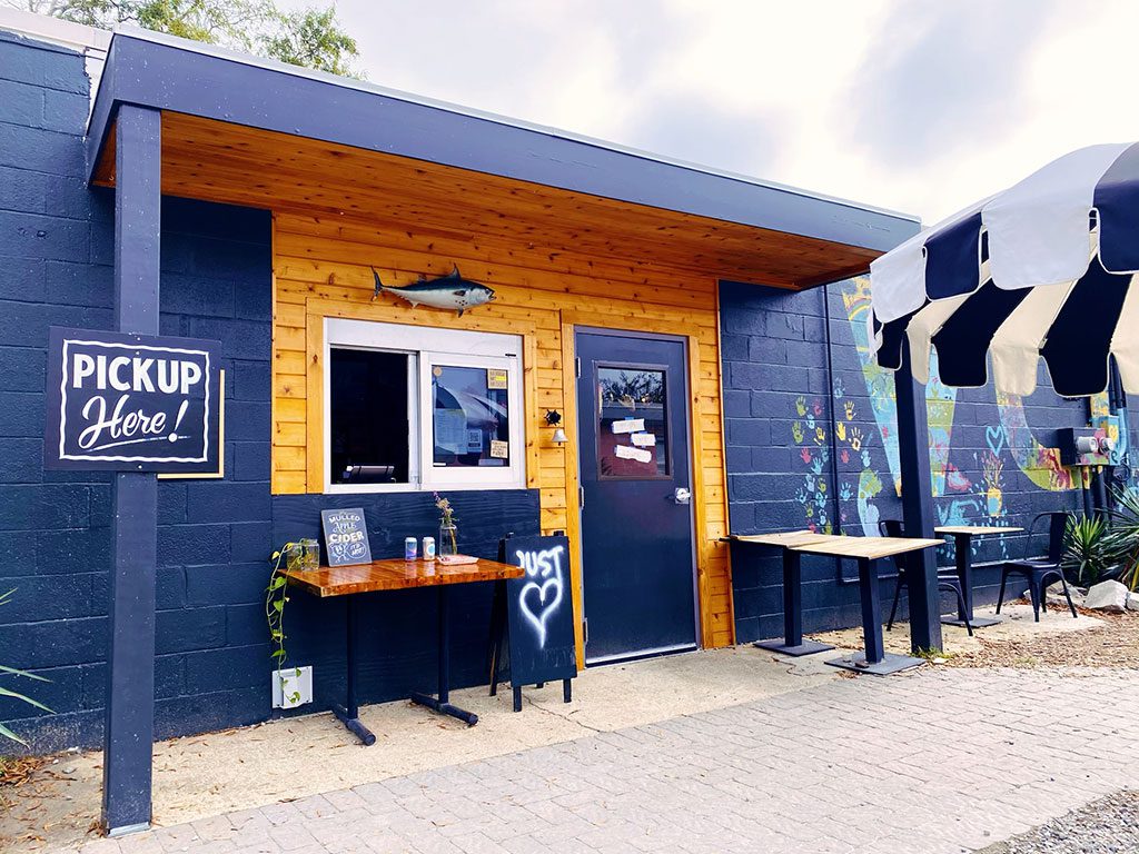 The exterior of Three Ships Coffee Roasters with wooden accents and a "PICKUP Here!" sign, potentially one of the unique coffee shops in Virginia Beach VA offering takeout, is shown with outdoor tables and a whimsical mural.