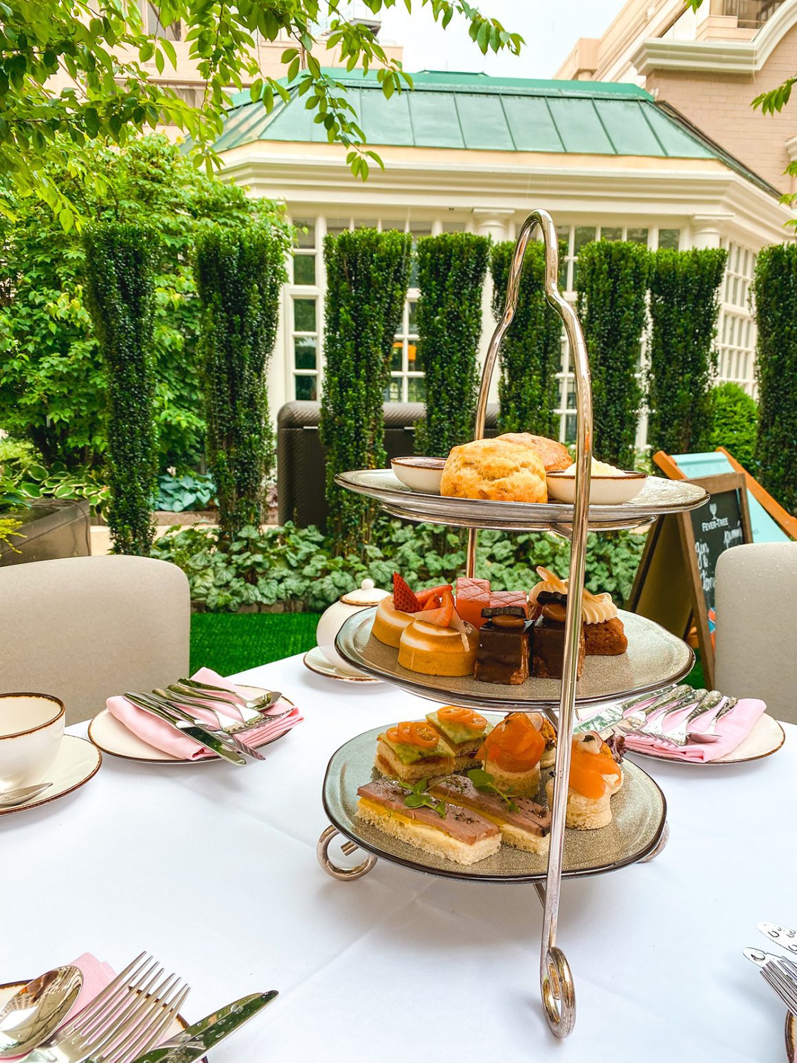 A beautifully set table at the Fairmont, ready for afternoon tea Washington DC, features a tiered tray filled with scones, pastries, and sandwiches, set against a lush green garden backdrop. - credit Keryn Means of DCTravelMag.com and a DC travel expert